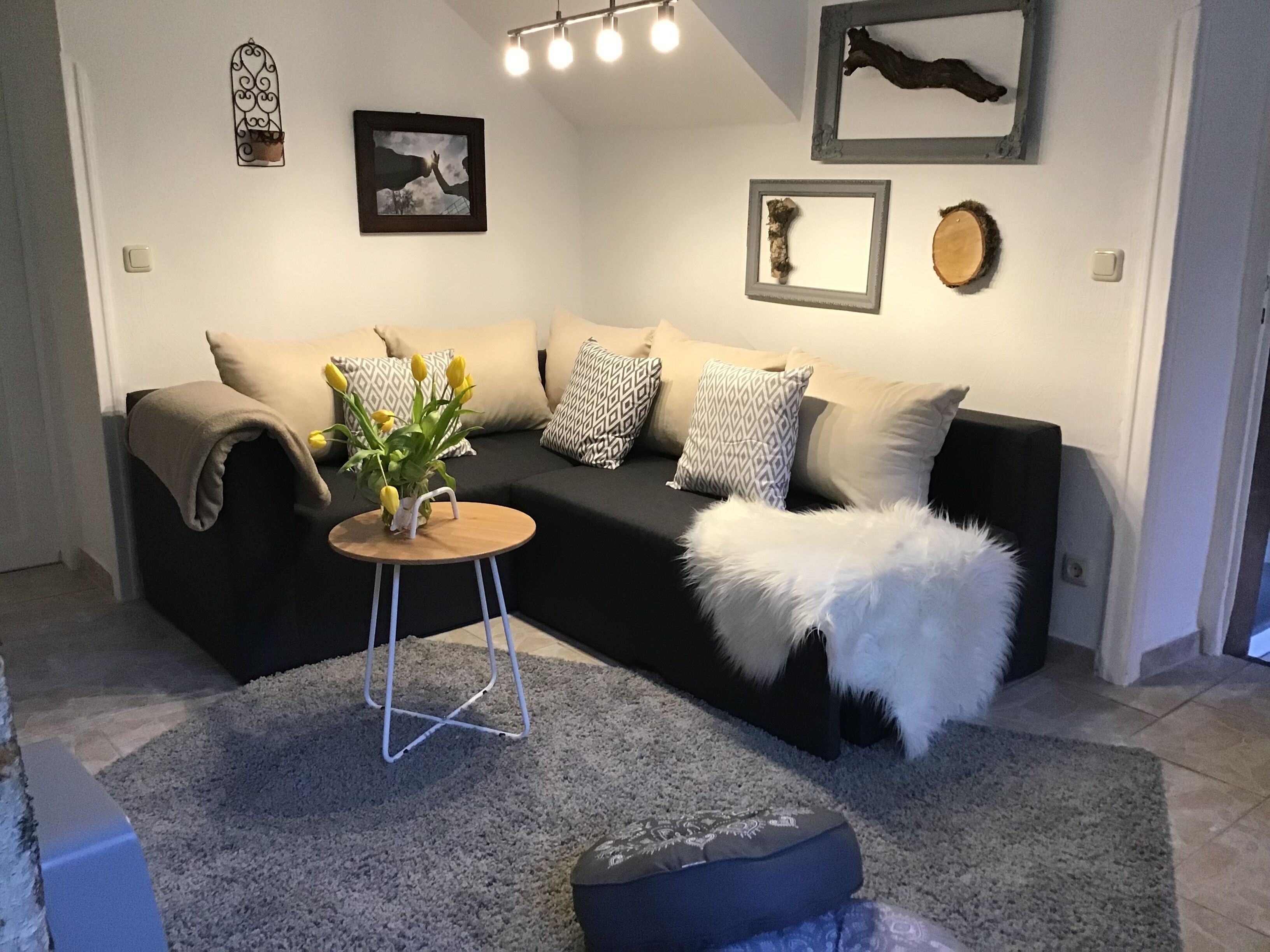 Un salon moderne avec un canapé noir, des coussins à motifs, une petite table d’appoint en bois sur laquelle est posé un vase rempli de fleurs jaunes, et un plaid blanc moelleux.