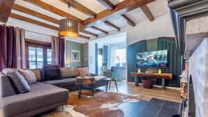 Ein modernes Wohnzimmer mit einer Eckcouch, einem Couchtisch aus Holz und einem Kuhfellteppich. An einer grünen Wand ist ein Fernseher angebracht, und rechts ist ein Kamin zu sehen.
