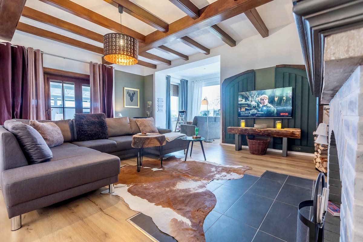 A modern living room with a sectional sofa, a wooden coffee table, and a cowhide rug. There is a television mounted on a green wall, and a fireplace is visible on the right.