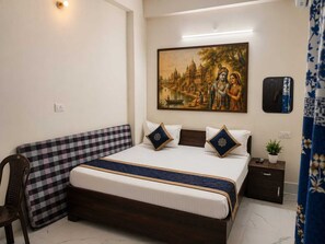 A hotel room with a bed, a checkered headboard, a painting of a temple scene, a nightstand with a plant, and a mirror. - Gopal kung (Mathura)