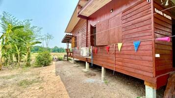A wooden house with a porch, surrounded by greenery and a dirt path.