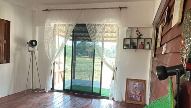 A room with a wooden floor, a sliding glass door leading to a garden, a tripod, and a framed picture on the wall. - Baan Praew Farm stay (Cottage) (Soem Ngam)
