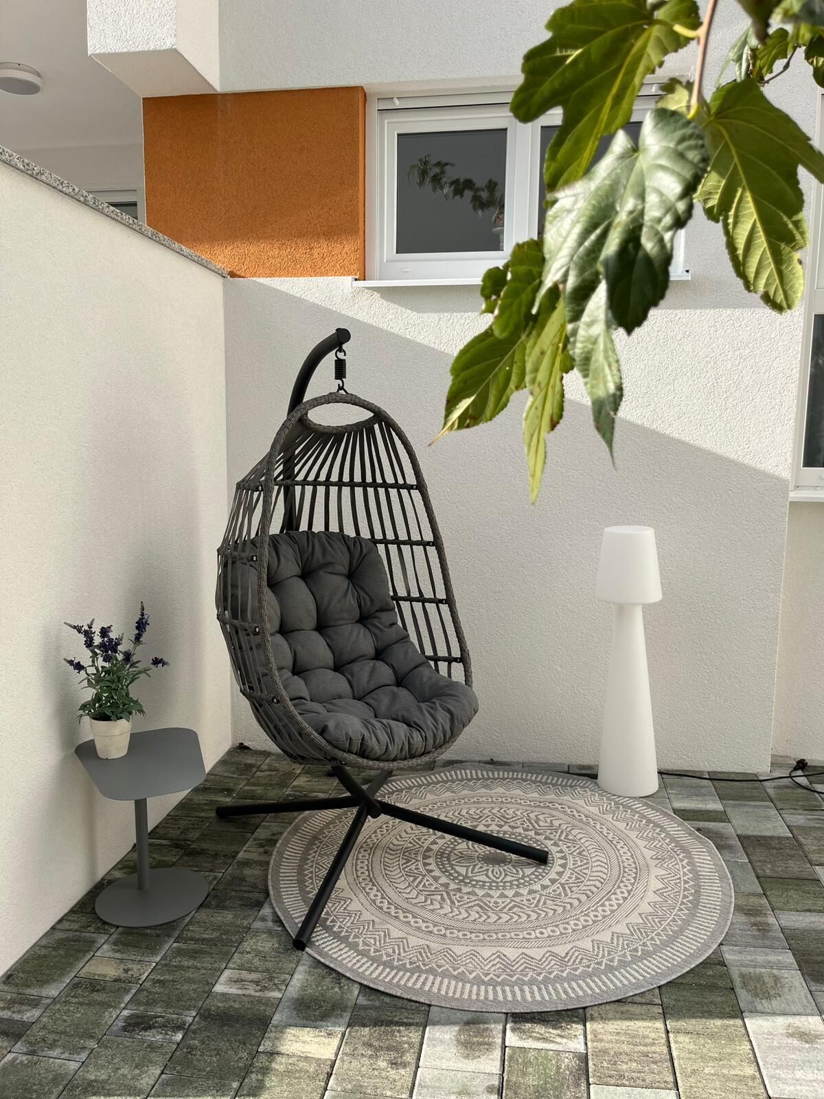 Una silla colgante con cojines, una planta pequeña en maceta, una lámpara de pie blanca y una alfombra estampada sobre un suelo de baldosas.