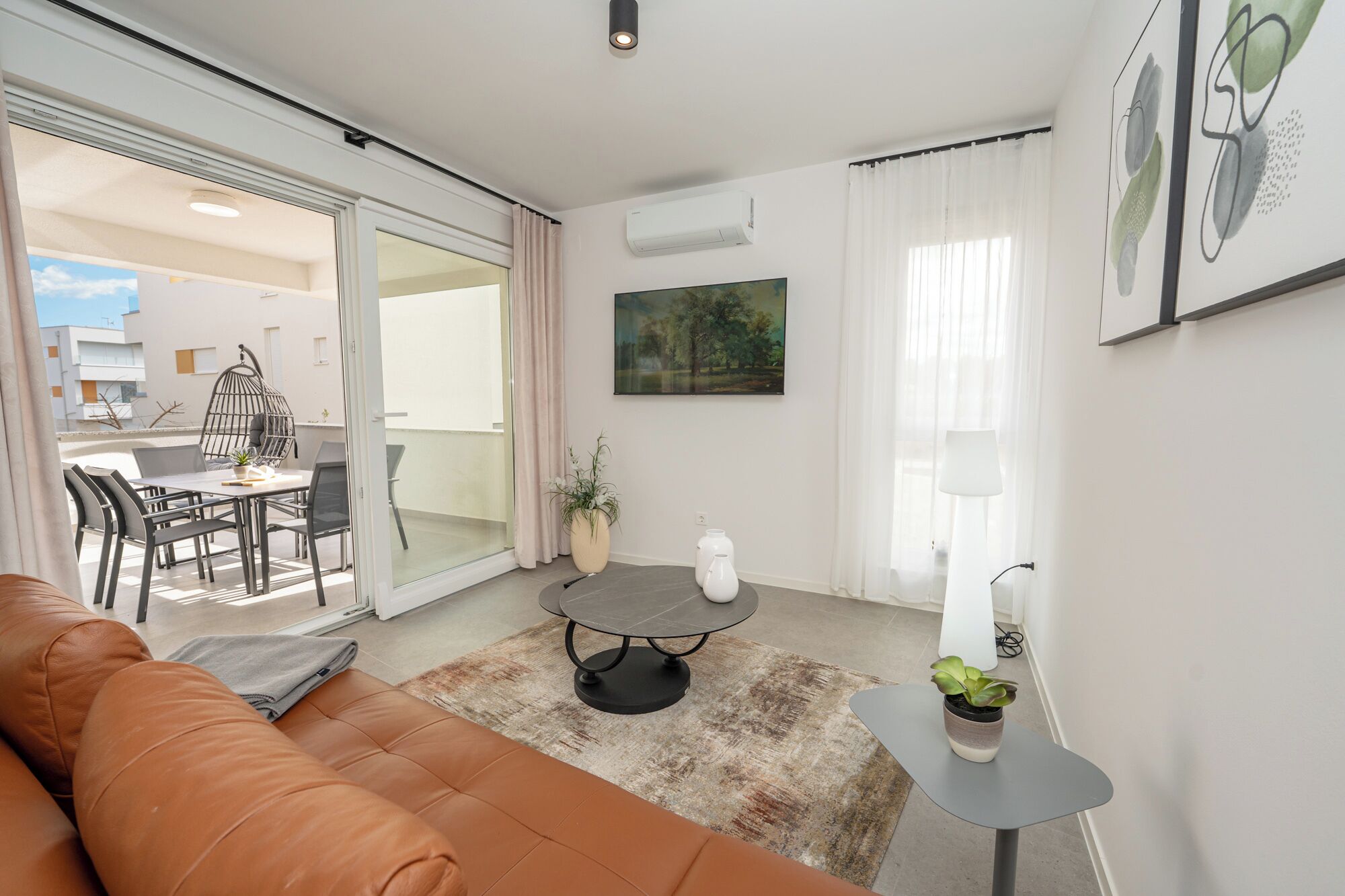 Ein modernes Wohnzimmer mit einem Ledersofa, einem Esstisch mit Stühlen, einem kleinen runden Tisch mit einer Vase und einem großen Fenster mit Vorhängen.
