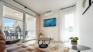 Een moderne woonkamer met een leren bank, een ronde salontafel en een eethoek met stoelen en een tafel. Er hangt een schilderij aan de muur, staat een potplant en is er een airconditioner aan het plafond bevestigd.