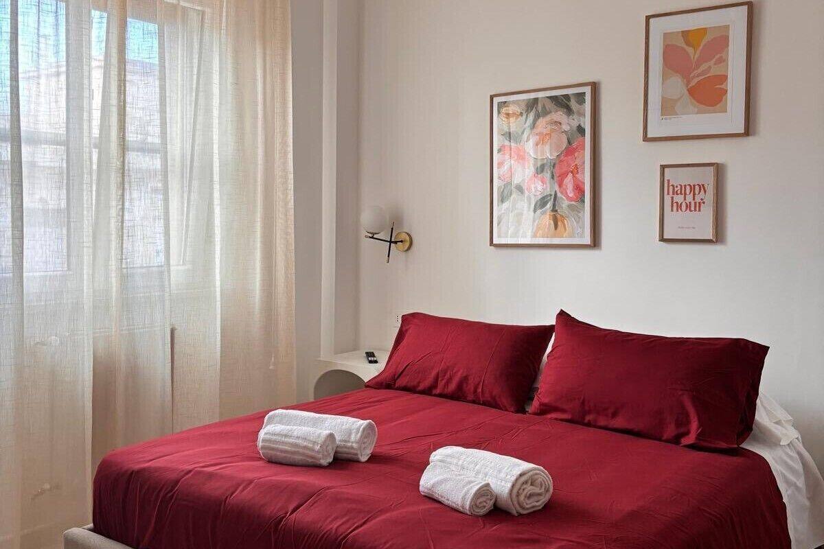 Een net opgemaakt bed met rode beddengoed en twee opgerolde handdoeken, een wandklok en twee ingelijste kunstwerken.