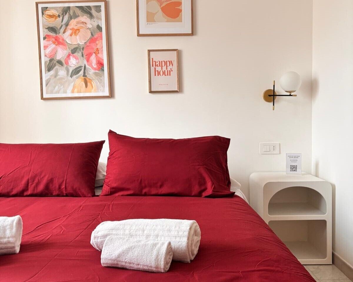 A hotel room with a red bedspread, two pillows, a white bedside table with a lamp, and a framed floral painting on the wall.