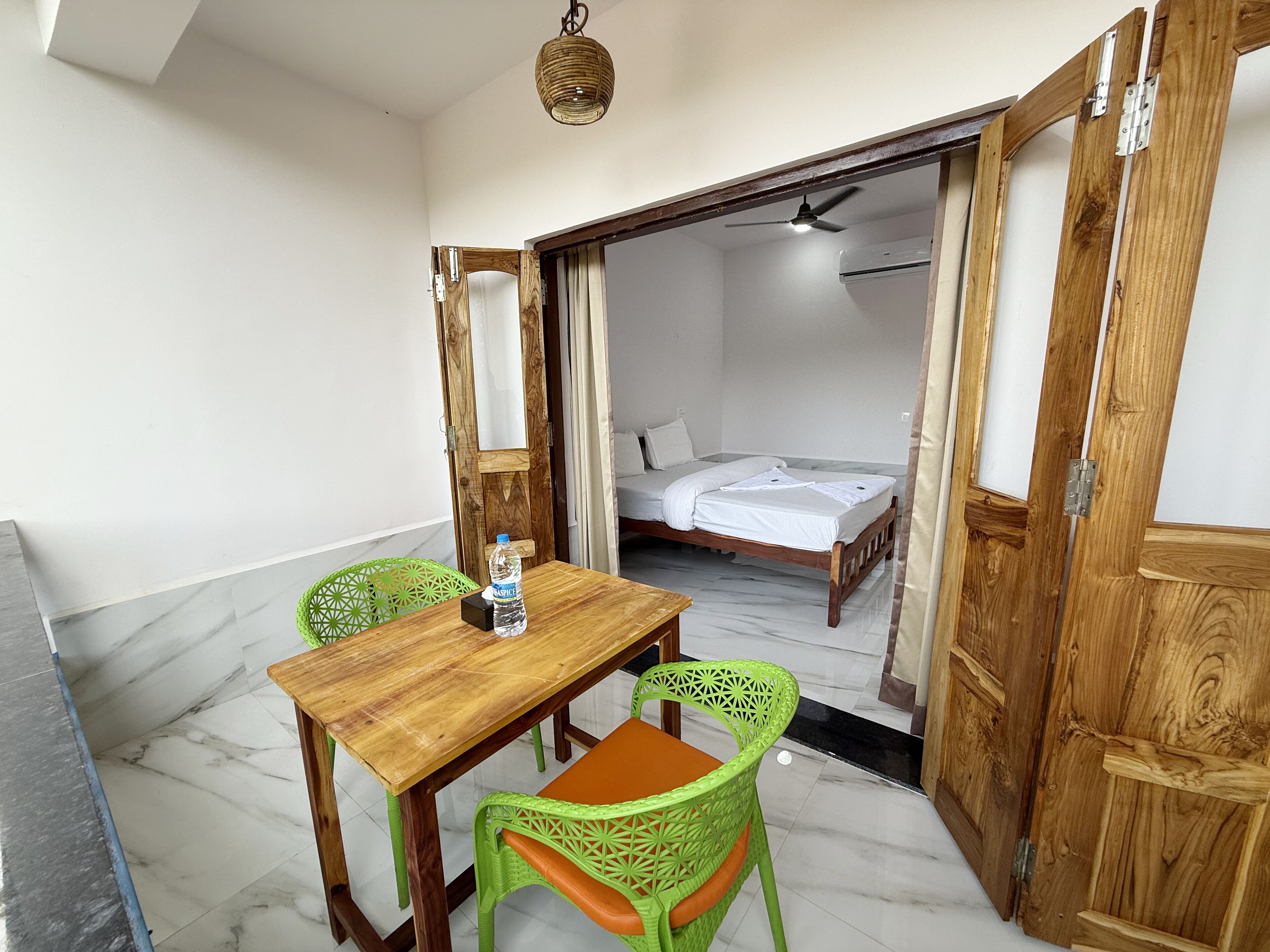 A room with a wooden table and two green chairs, a bed in the adjacent room, and a ceiling fan.