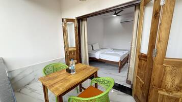 A room with a wooden table and two green chairs, a bed in the adjacent room, and a ceiling fan.
