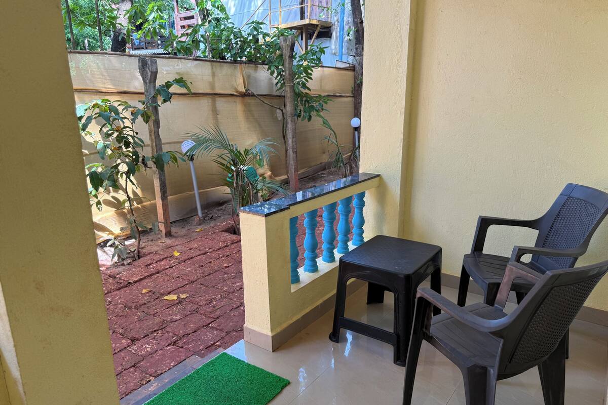 A small balcony with a table and two chairs, a green mat, and a view of a garden with plants and a fence.