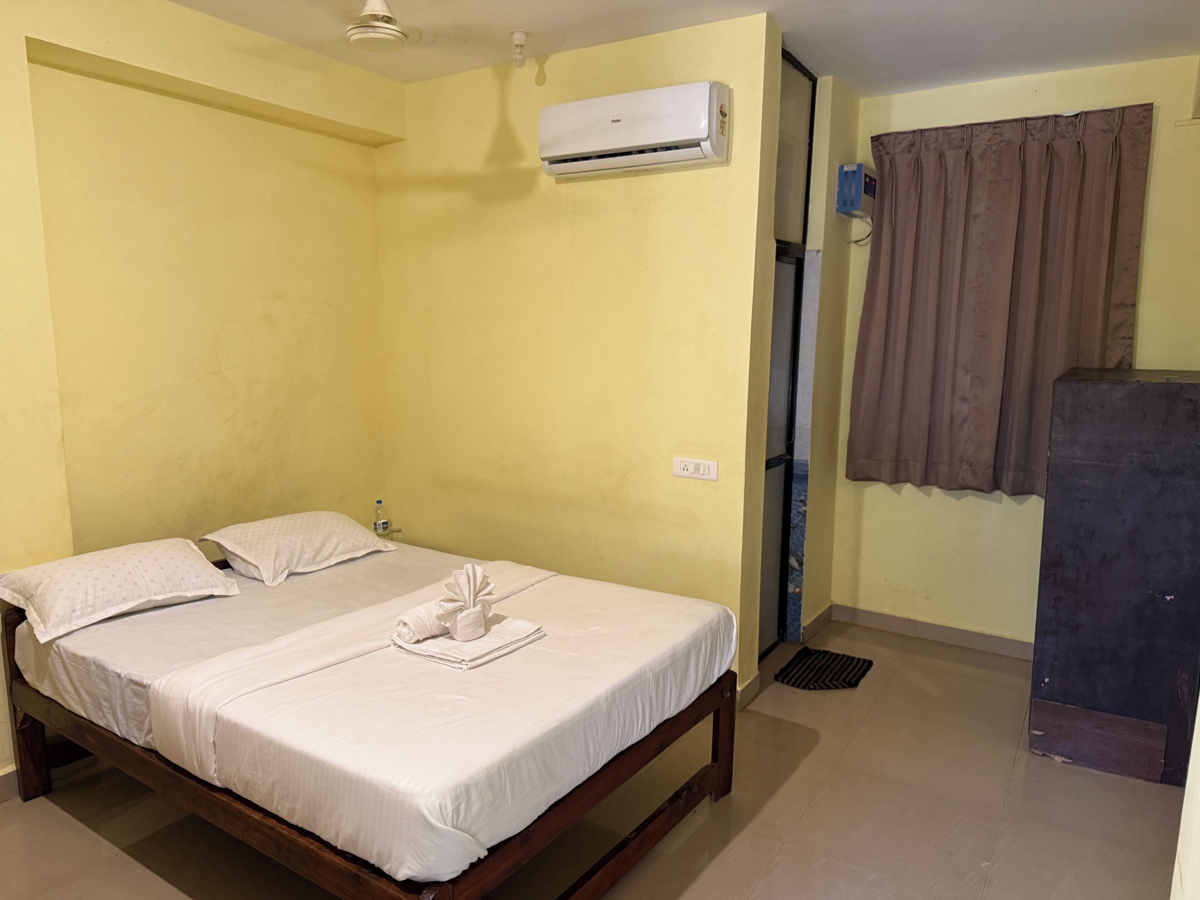 A single bed with a white mattress and pillows, a wooden frame, an air conditioning unit on the wall, a brown curtain, and a small table with a bottle.
