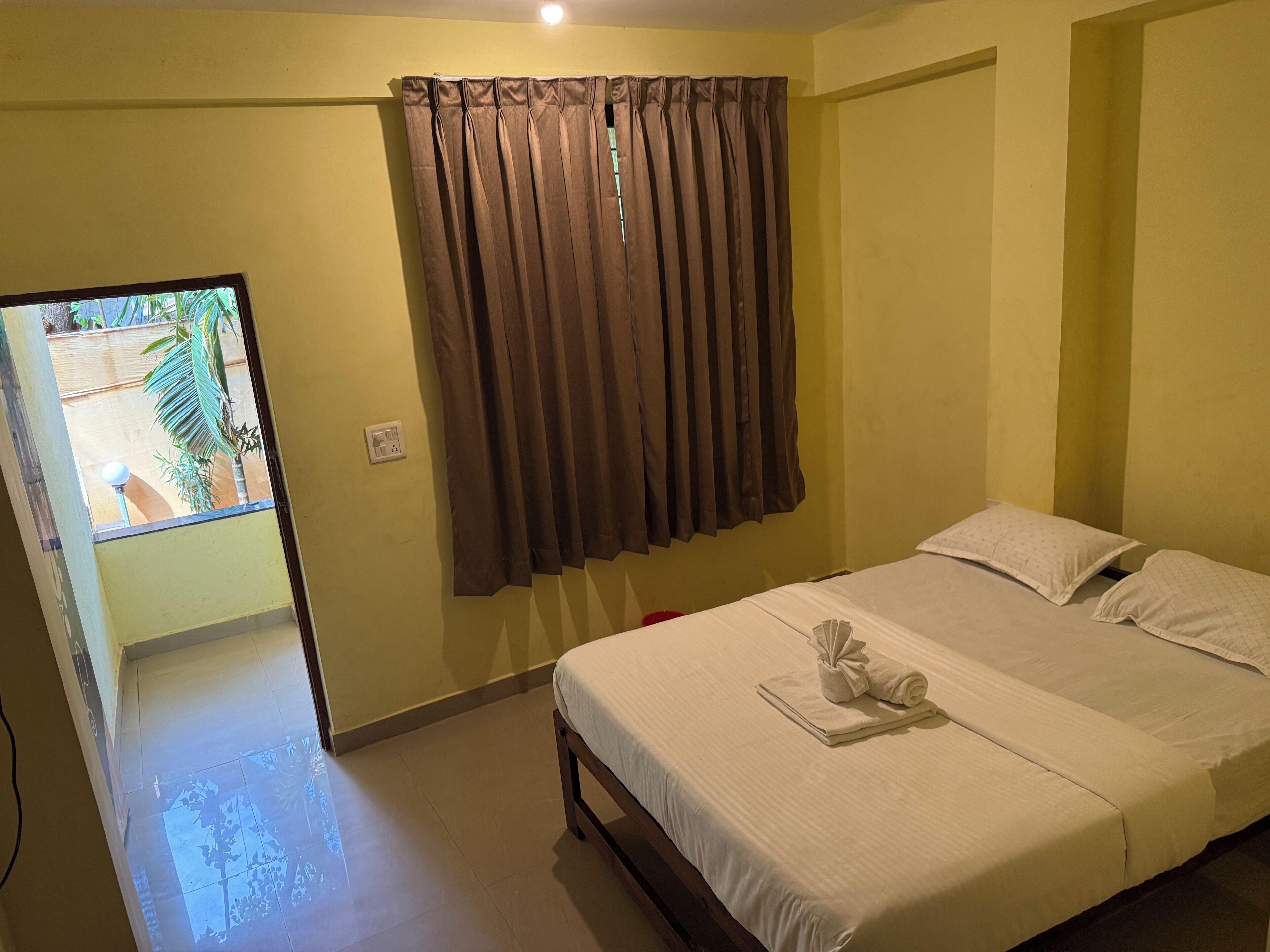 A hotel room with a bed, a window with curtains, a small table with a towel, and a view of the outside.
