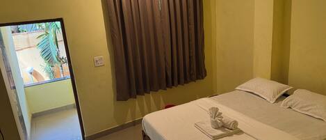 A hotel room with a bed, a window with curtains, a small table with a towel, and a view of the outside.