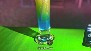 Un vaso alto con un efecto de color degradado, lleno de una bebida y adornado con hierbas, sobre una superficie verde con un fondo desenfocado.