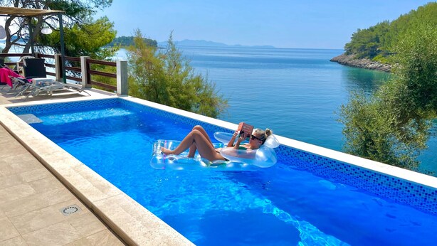 A pool with clear blue water, surrounded by greenery and a view of the sea. - Waterfront House Arnet by Villas Guide (Blato)