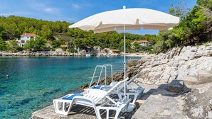 A coastal scene with clear water, a white umbrella, and lounge chairs on a rocky shore. - Waterfront House Arnet by Villas Guide (Blato)