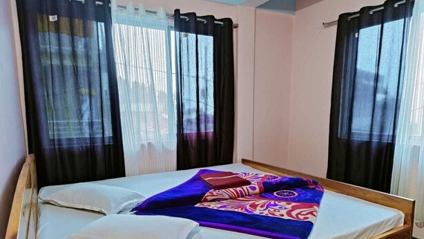 A single bed with a white mattress and a colorful blanket, two black curtains, and a window with a view of the outside. - Sherpa Residency (Kalimpong)