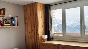 A bedroom with a bed, wooden wardrobe, bookshelves, a desk, and a window with a mountain view. - Alpine Escape (Huez)