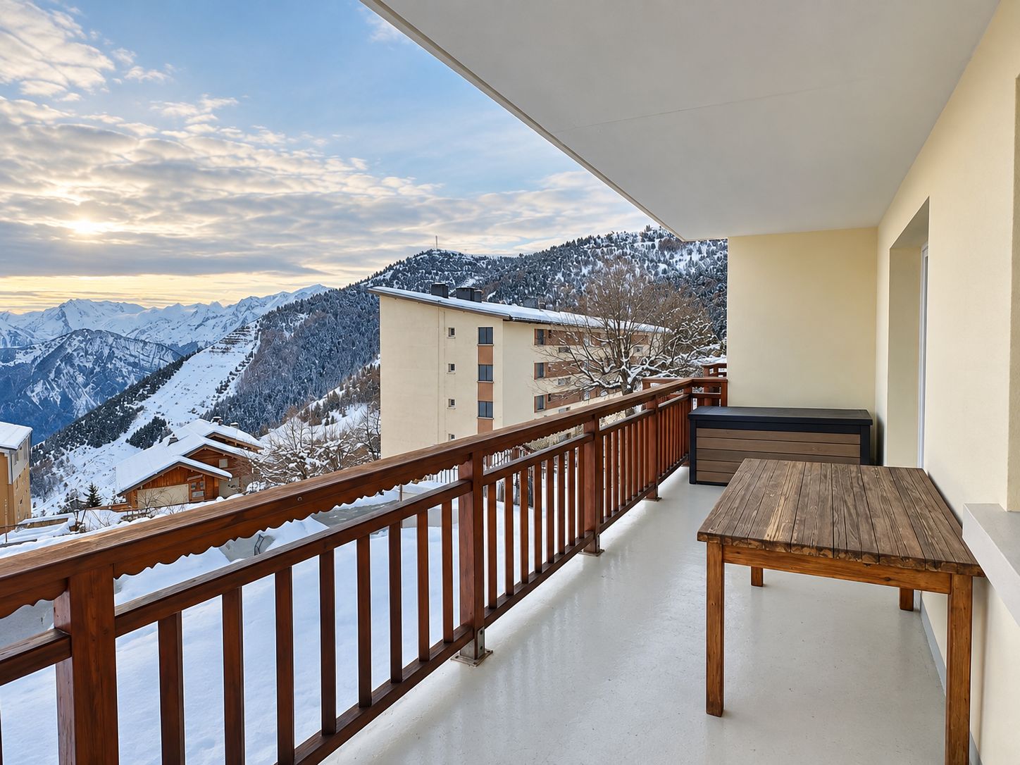 A balcony with a wooden railing, a wooden table, and a view of snow-covered mountains and buildings.