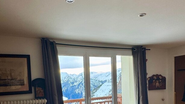 A living room with a large window, a radiator, a sofa, and a small round ottoman. - Alpine Escape (Huez)