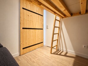 A room with wooden flooring, a wooden door with black horizontal bars, a ladder leaning against the wall, and a white wall with a power outlet. - Escape from the Alps (Huez)