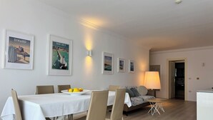 Un comedor con una mesa blanca, sillas y un plato de fruta. Hay cuadros en la pared y una lámpara al fondo.