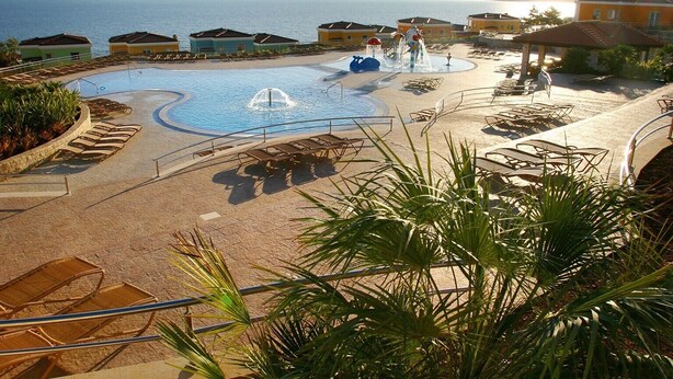 Zona de piscina en un resort con fuente de agua, sillas de descanso y vista al mar.