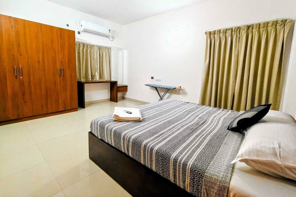 A bedroom with a bed, a wardrobe, an ironing board, a ceiling fan, and a window with curtains.