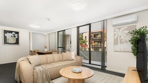 Ein modernes Wohnzimmer mit einem beigen Sofa, einem runden Couchtisch aus Holz und einem Fernseher auf einer Holzablage.