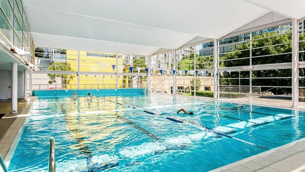 Ein Hallenbad mit deutlich abgegrenzten Bahnen, umgeben von einem modernen Gebäude mit großen Fenstern und Balkonen.