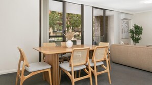 Ein modernes Wohnzimmer mit einem Holzesstisch und -stühlen, einem beigen Sofa und einem großen Fenster mit Blick auf Grünflächen und Gebäude.
