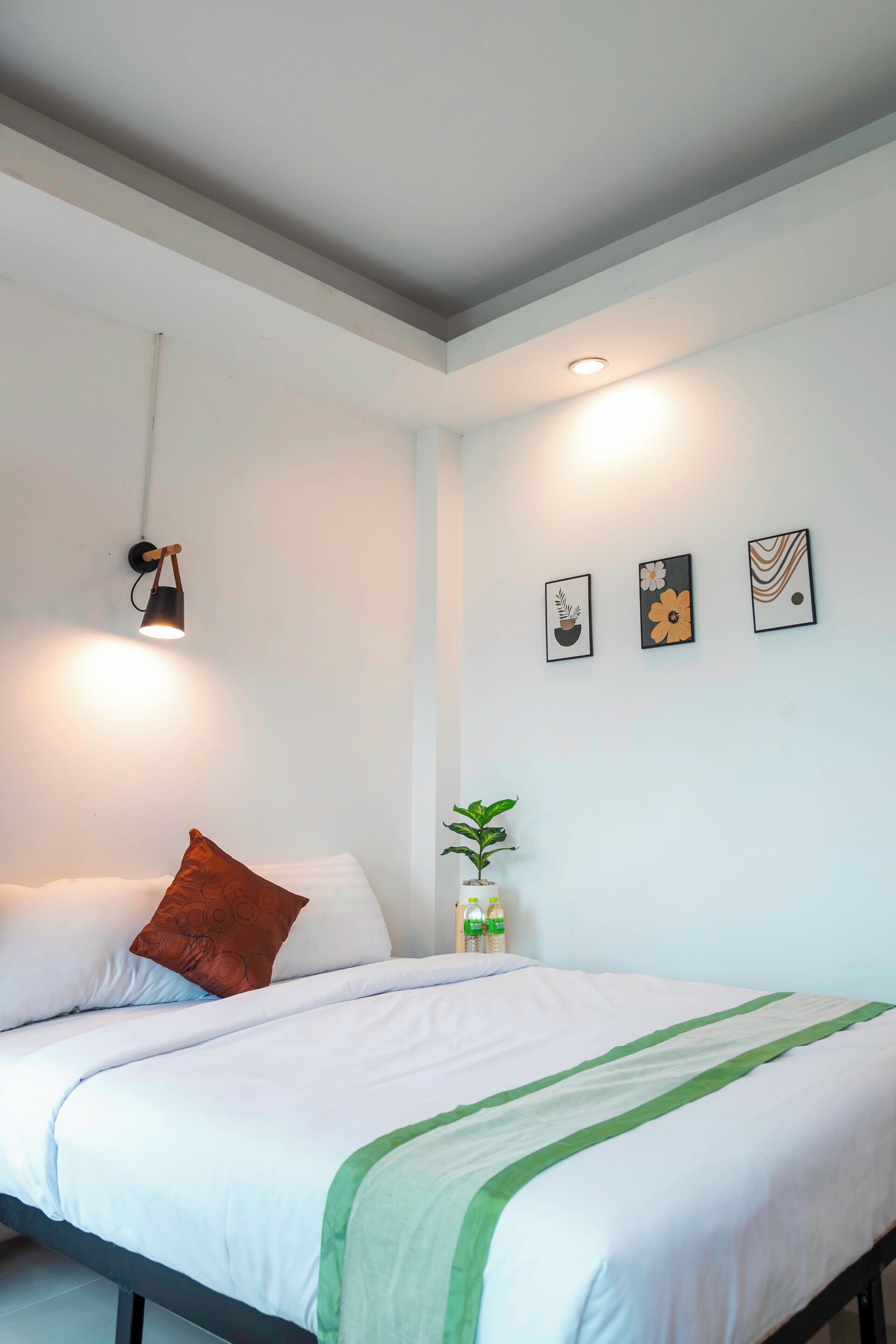 Cama bem arrumada com roupa de cama branca e detalhes verdes, uma luminária de parede e três quadros emoldurados.