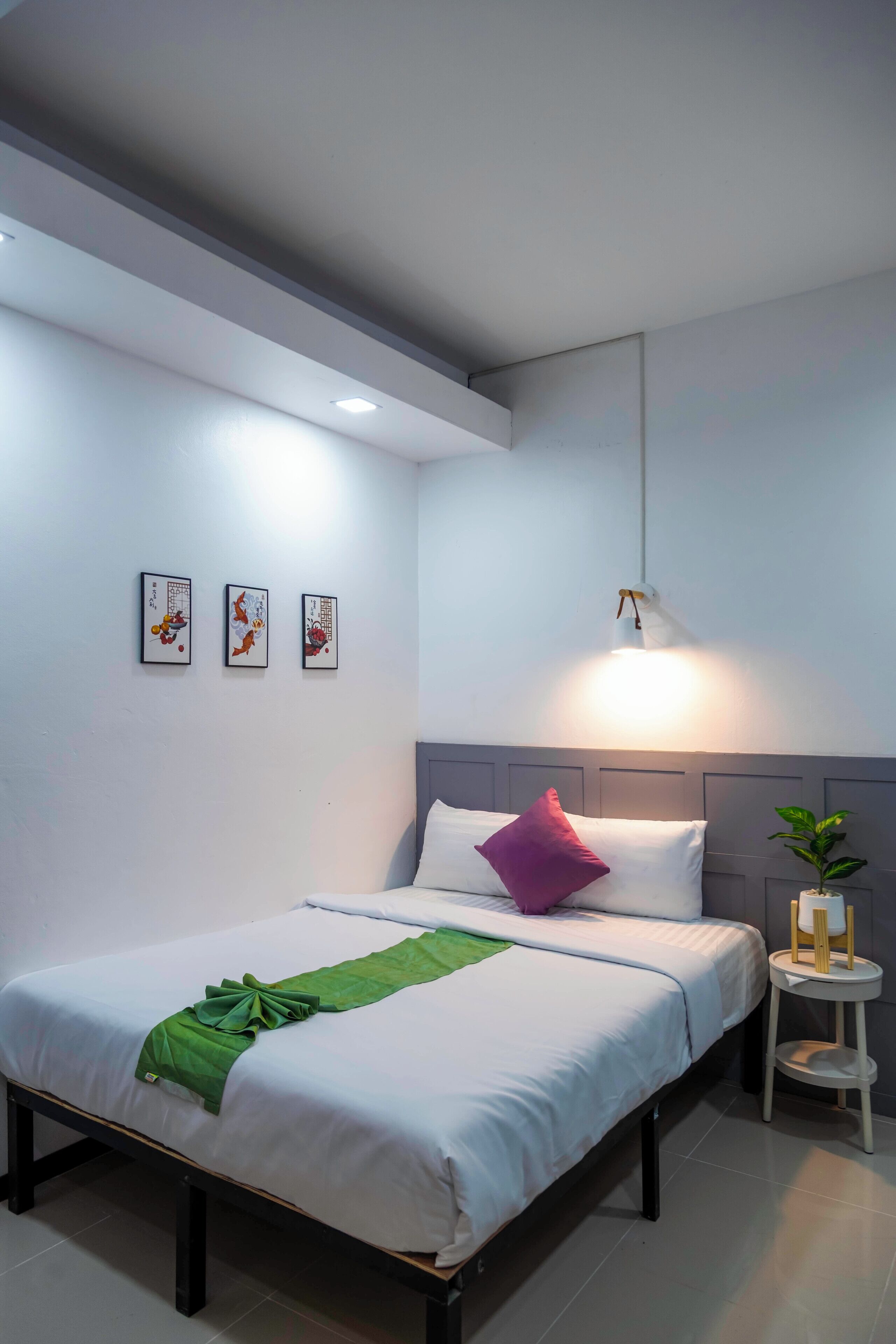Una cama bien hecha con una toalla verde, un almohajar blanco y un almohajar morado. En la pared hay tres cuadros enmarcados, una pequeña mesa blanca con una planta y una lámpara colgante.
