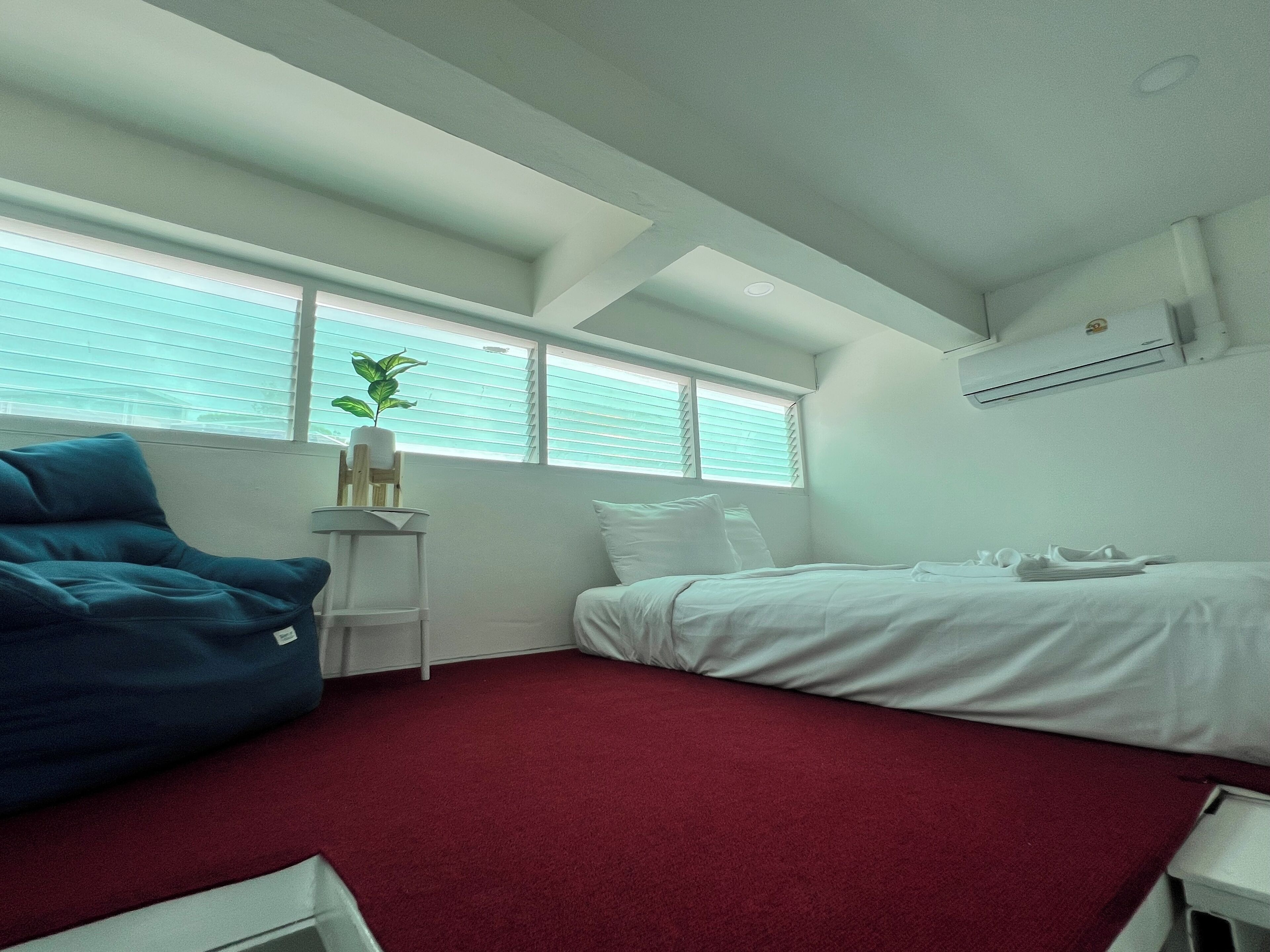 A bedroom with a bed, a blue chair, a small table with a plant, and a window with blinds.