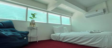 A bedroom with a bed, a blue chair, a small table with a plant, and a window with blinds.