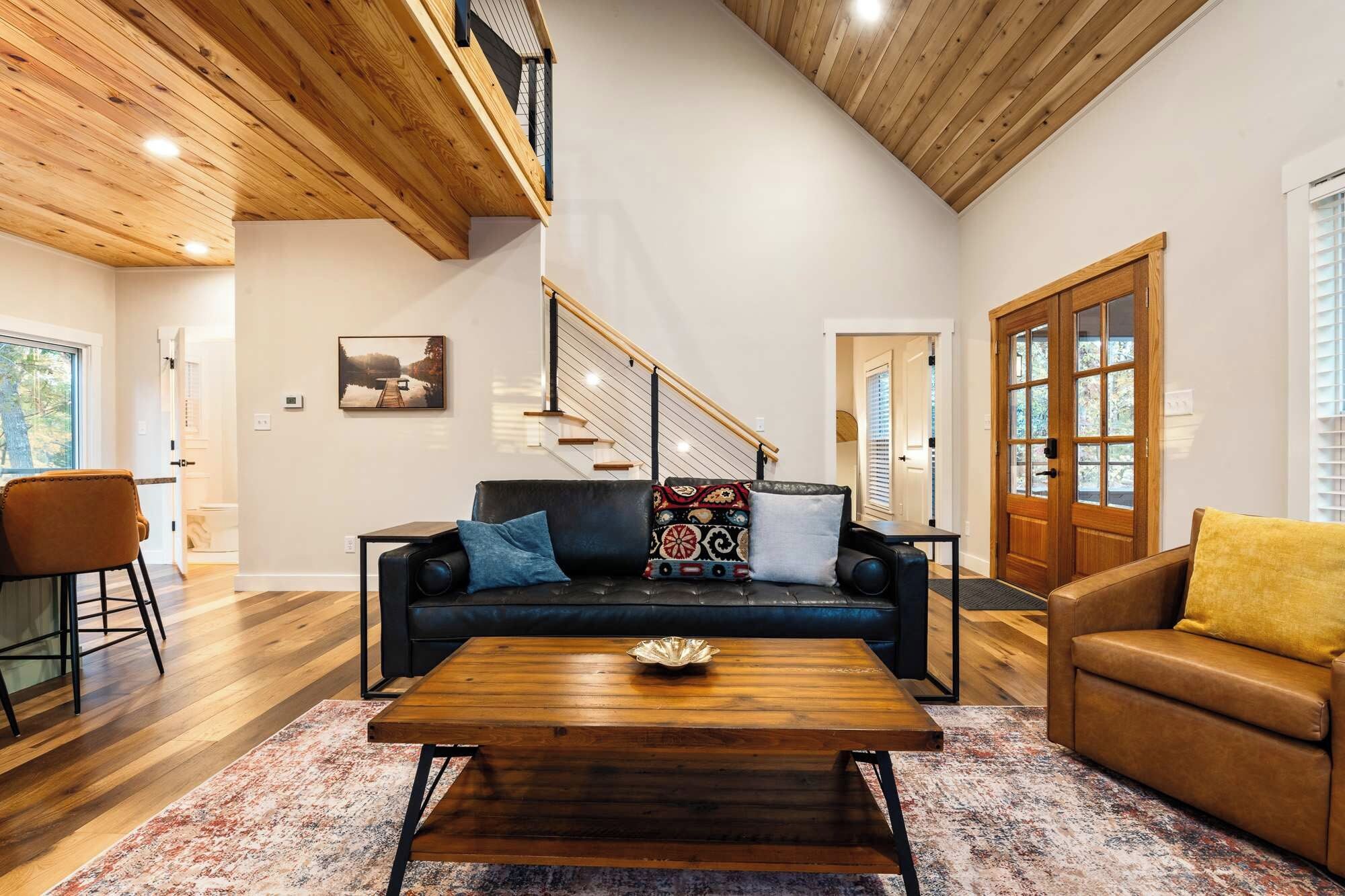 Un vaste salon doté d’un haut plafond, d’un escalier en bois, d’un canapé en cuir noir agrémenté de coussins à motifs, d’une table basse en bois et d’un fauteuil jaune.