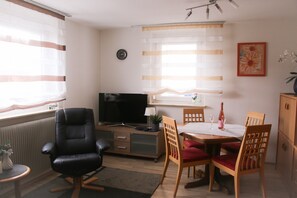 Ein Wohnzimmer mit Fernseher, einem Esstisch mit Stühlen, einer Couch, einem kleinen Tisch mit Lampe und einer Heizung.