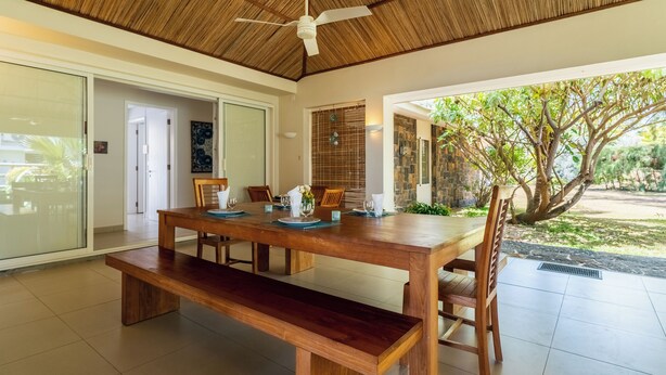 Comedor con mesa de madera dispuesta para cuatro personas, ventilador de techo y vista a un jardín con árboles y un muro de piedra.