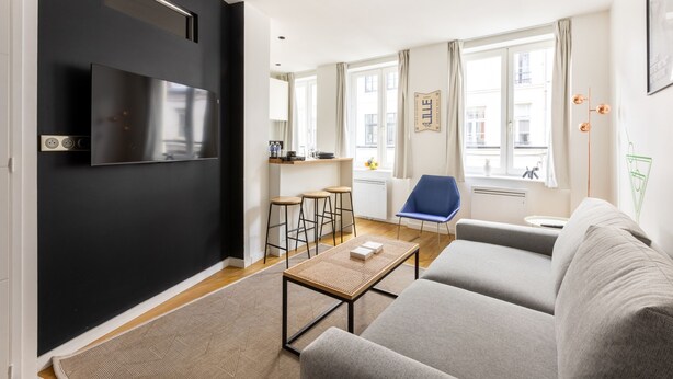 Un salon moderne comprenant un canapé, une table basse, un téléviseur fixé au mur et un coin cuisine avec des tabourets.