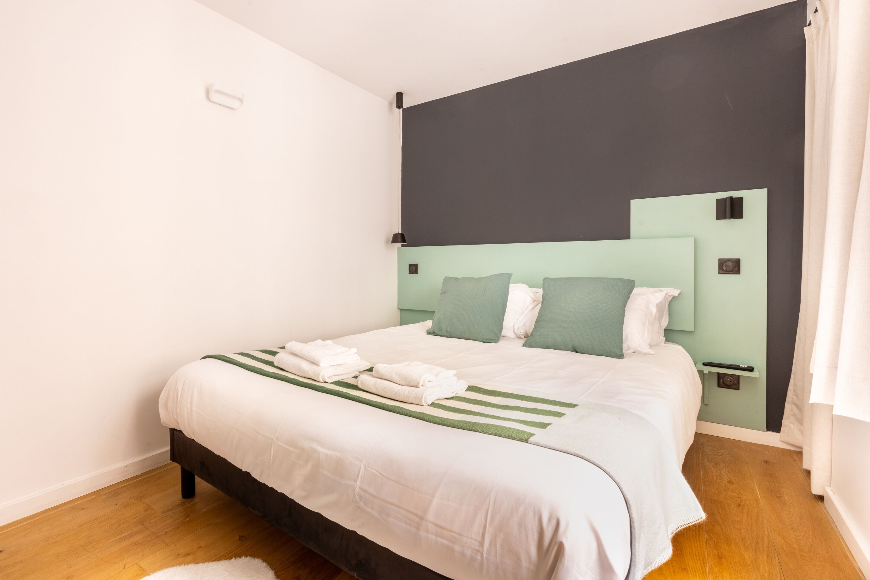 Een net opgemaakt bed met witte lakens en groene accenten, een donker hoofdbord en een klein nachtkastje met een lamp.