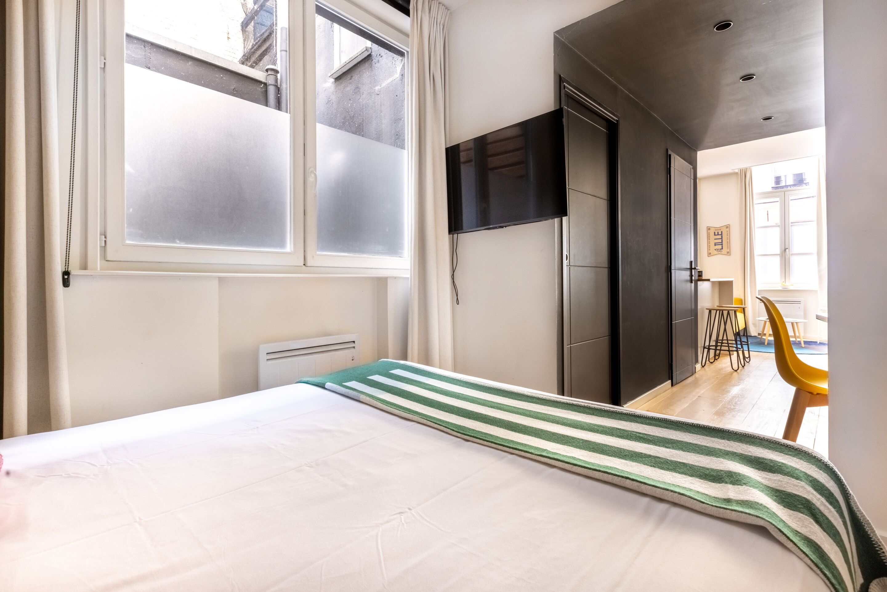A hotel room with a bed, a TV mounted on the wall, a window with frosted glass, and a view of the outside.