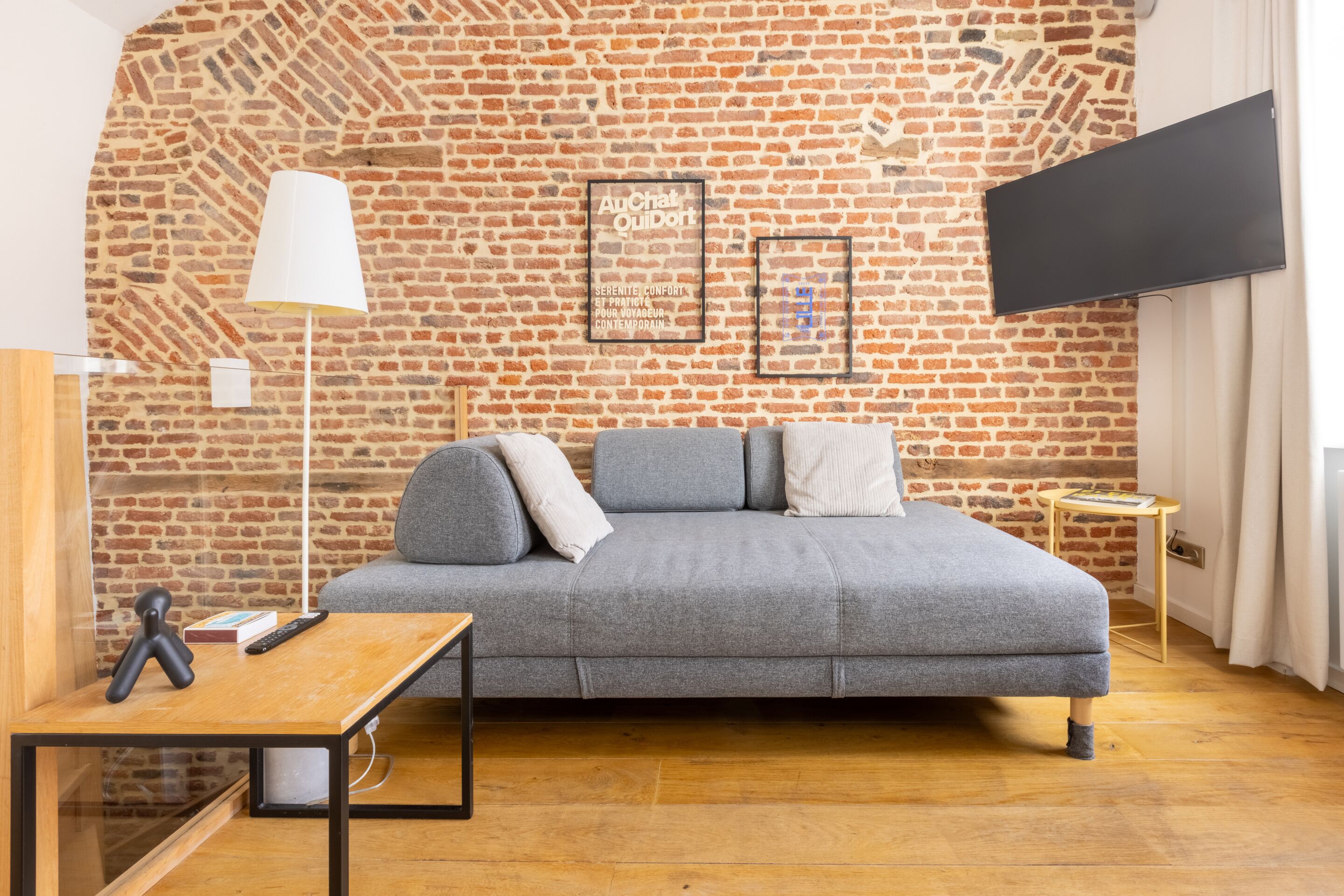 Ein gemütliches Zimmer mit Backsteinwand, einer grauen Couch, einem kleinen Holztisch und einem an der Wand befestigten Fernseher.