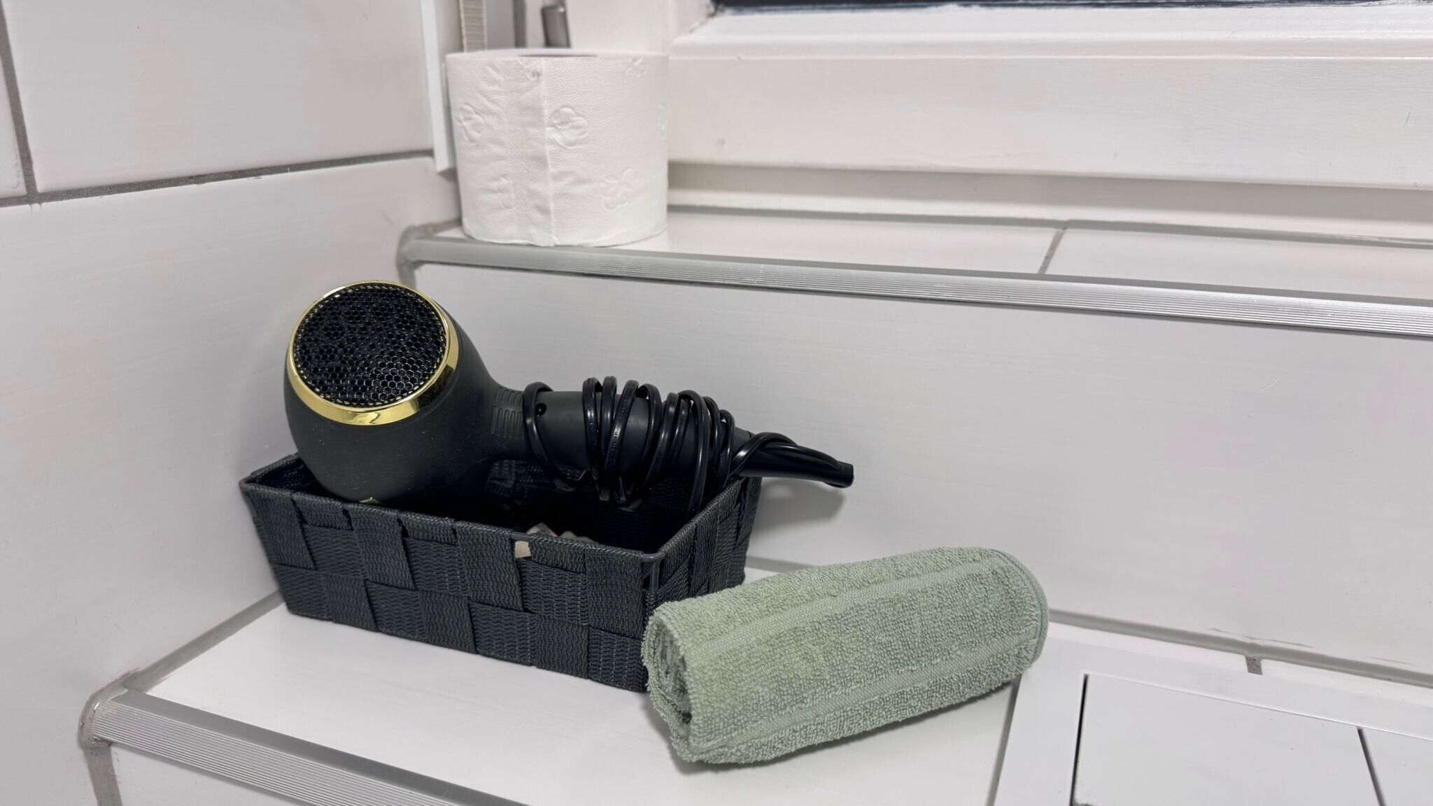 A bathroom with a towel rack, a hairdryer, and a basket containing a towel and headphones.