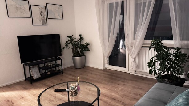 Sala de estar con televisor de pantalla plana, una mesa de centro de vidrio, sofá, planta en maceta y puerta corrediza de vidrio que da a un balcón.