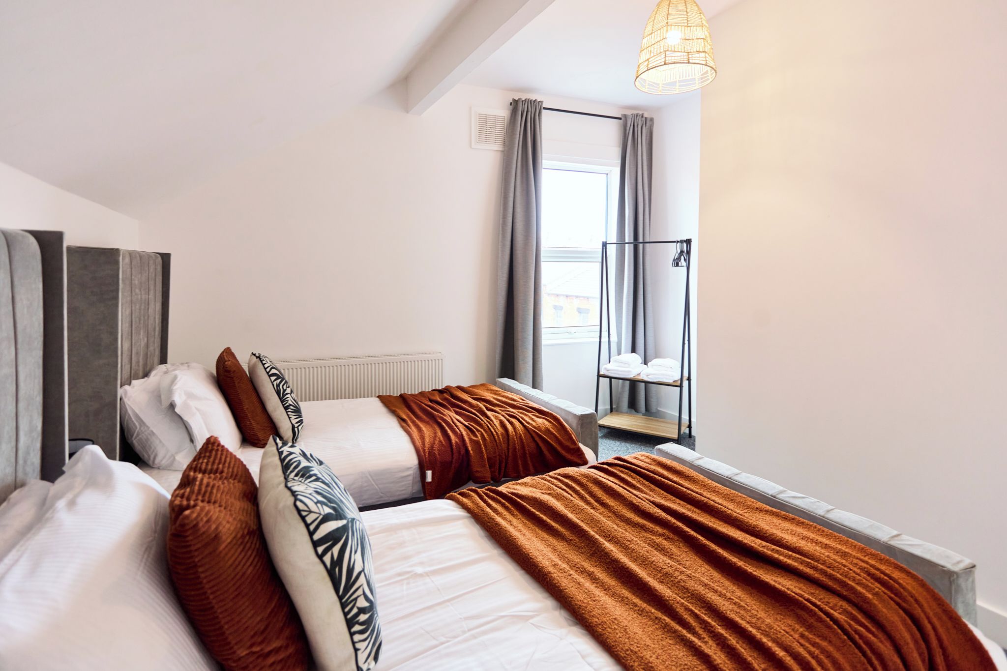 A modern hotel room with two beds, a window with curtains, a radiator, and a hanging light fixture.
