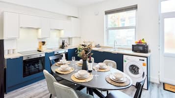 A modern kitchen with a dining area, white cabinets, a built-in oven, a washing machine, and a window with a view.