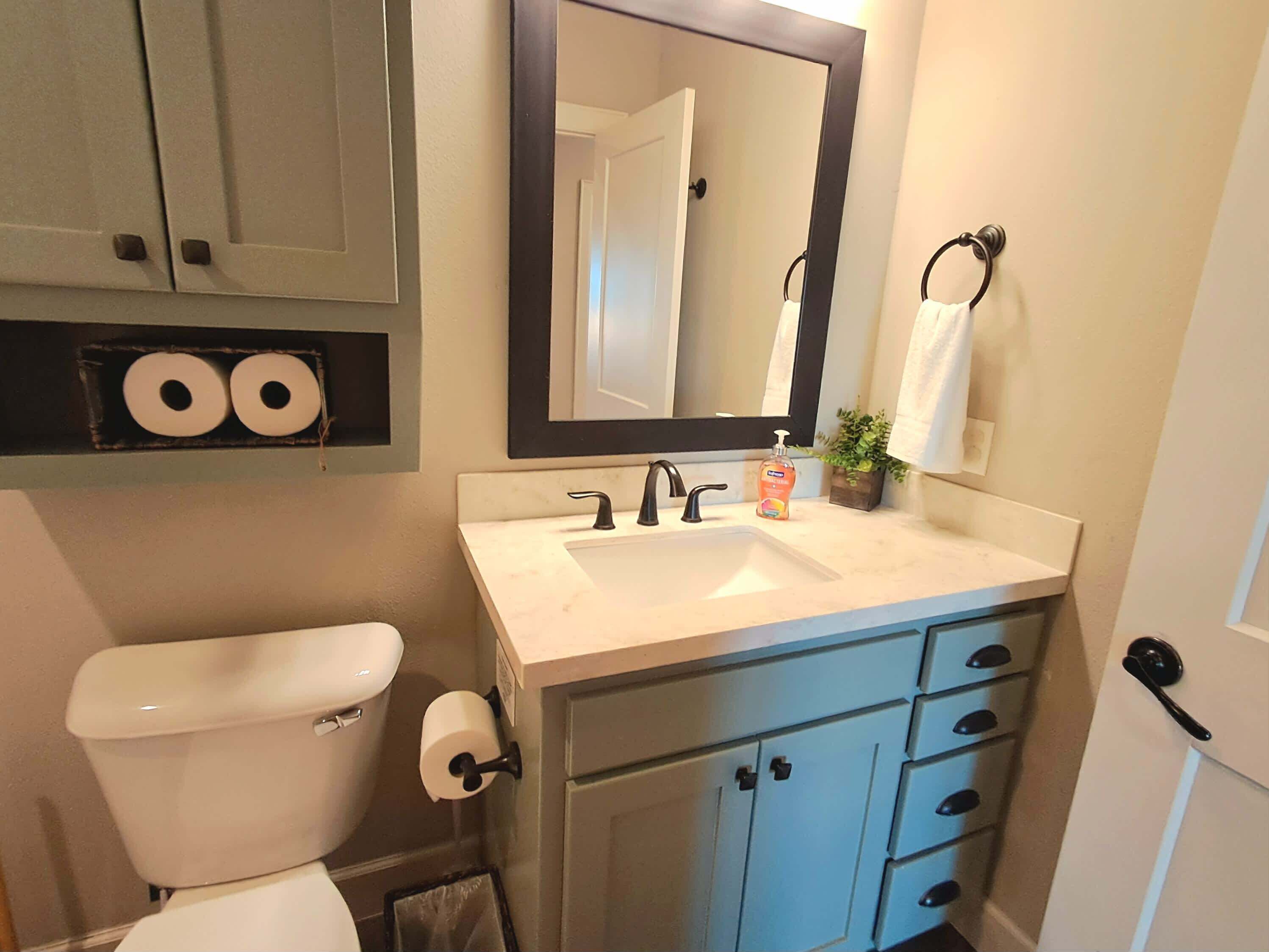 A bathroom with a toilet, sink, mirror, and cabinets.