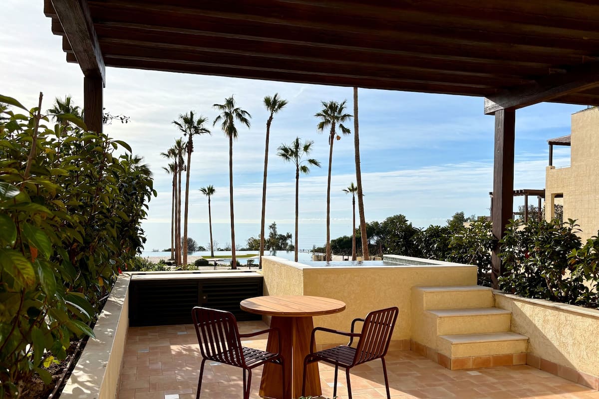 A covered outdoor seating area with a table and chairs, overlooking a pool and palm trees.