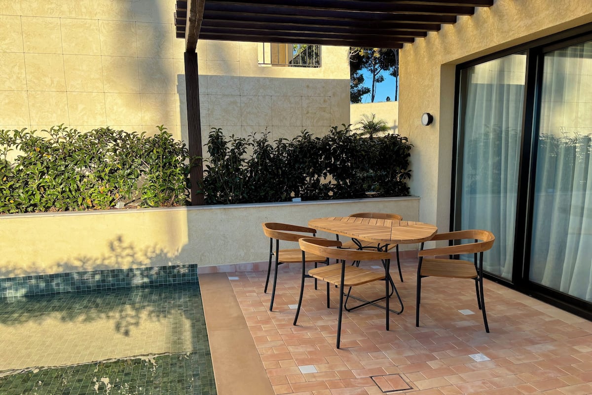A patio with a pool, wooden table and chairs, and a pergola.