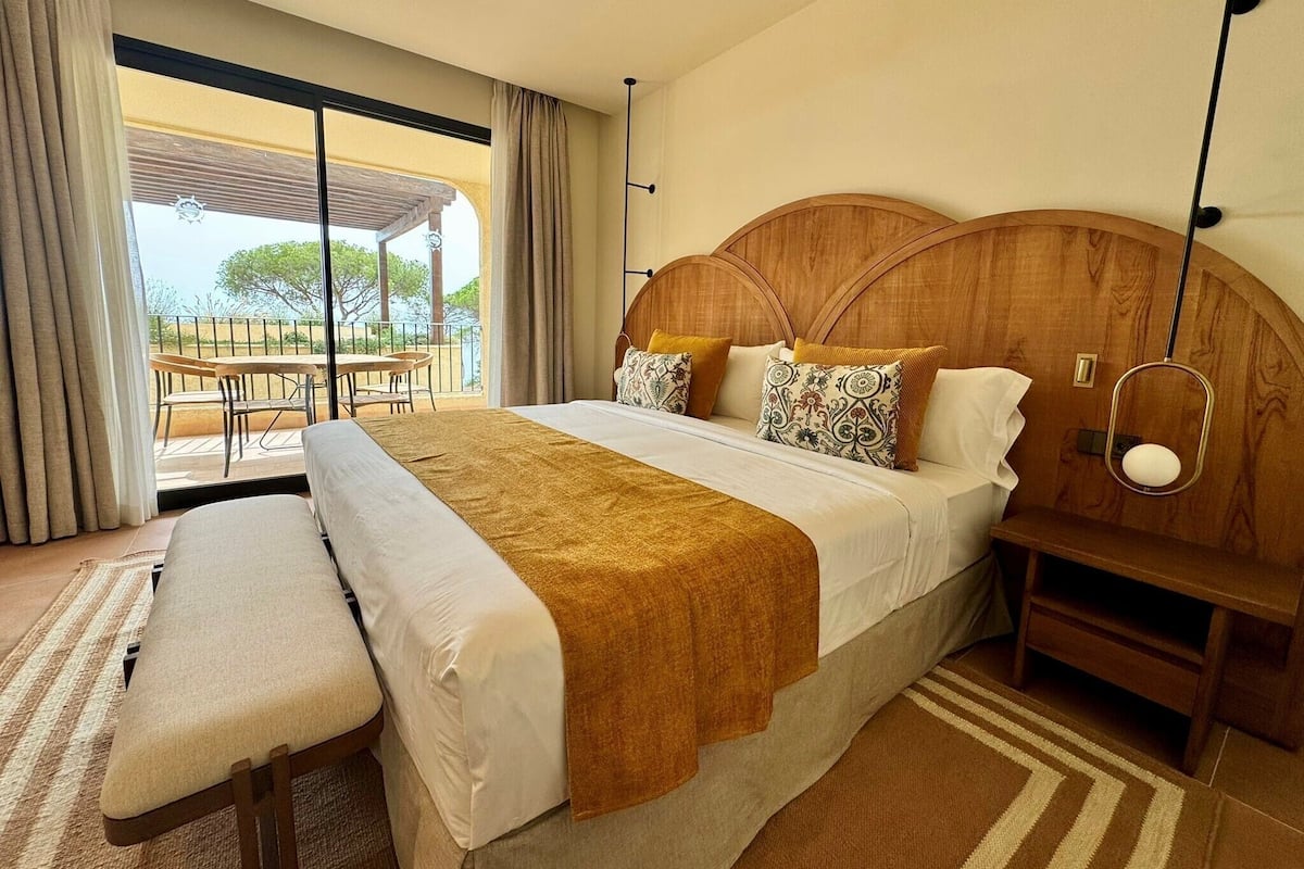 A spacious bedroom with a large bed, a wooden headboard, and a view of a balcony with outdoor furniture.